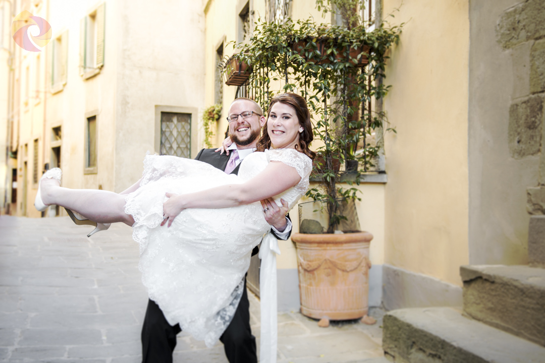 Lo sposo porta la sposa in braccio, entrambi ridono, in uno scorcio del borgo di Cortona