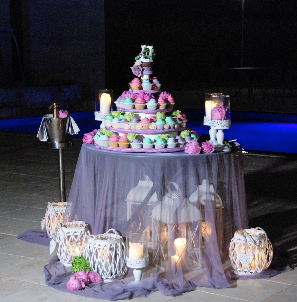 A wedding cake made of colorful cupcakes, surrounded by lamps and candles on a table with sheer tablecloth, with a lego cake topper