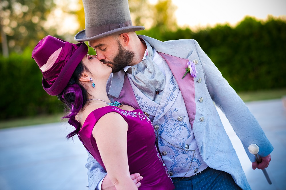 Spouses dressed in late victorian era fashion, sharing a passionate kiss in their top hats.