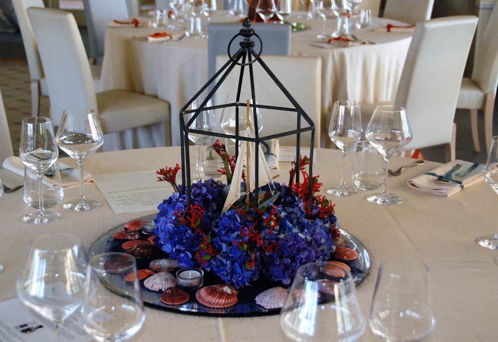 A centerpiece inspired by Moby Dick, by Herman Melville, with a sailboat inside an iron lamp frame, on a bed of seashells and blue and red flowers