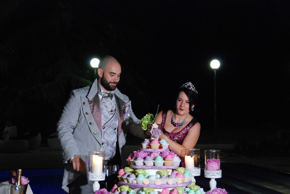 Cutting of the cake, with the victorian dressed spouses cutting the top of a cake made of colorful cupcakes, surrounded by candles