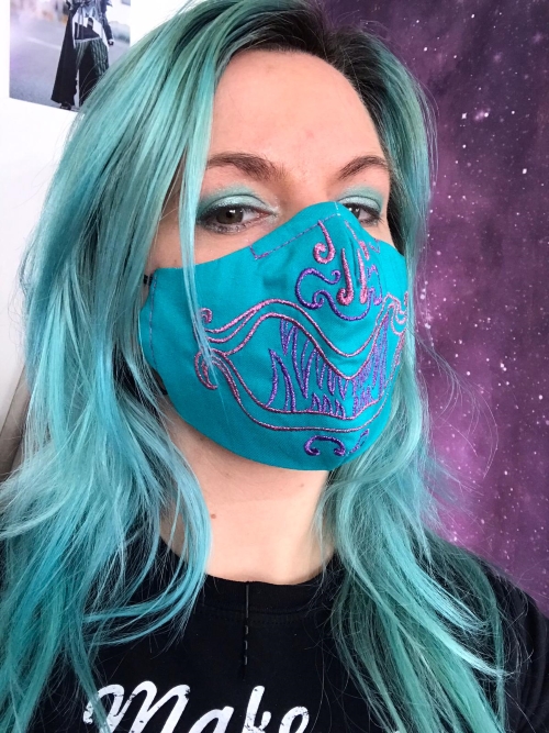 A girl with green/blue hair wearing a blue mask with a purple embroidery that resembles a monster.